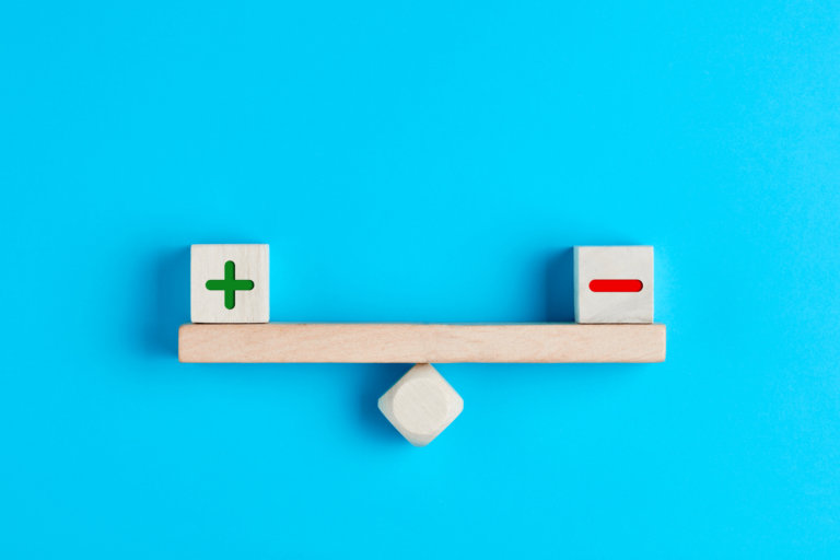 Plus and minus or positive and negative symbols on wooden blocks are in balance on a wooden seesaw. Blue background, flat lay view. Pros and cons equilibrium in decision making under uncertainity.
