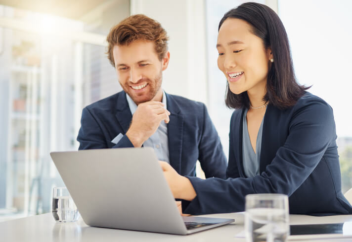 Two happy young diverse colleagues working together on a laptop in an office boardroom. Confident asian businesswoman explaining corporate plans and strategy to smiling caucasian businessman during a meeting