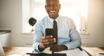 Smiling young African business executive sending text messages on his cellphone while sitting at his desk in an office