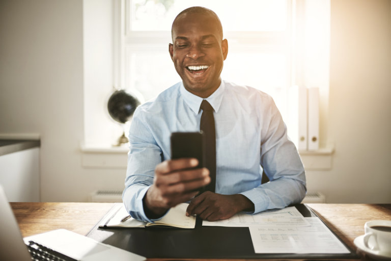 Smiling young African business executive sending text messages on his cellphone while sitting at his desk in an office