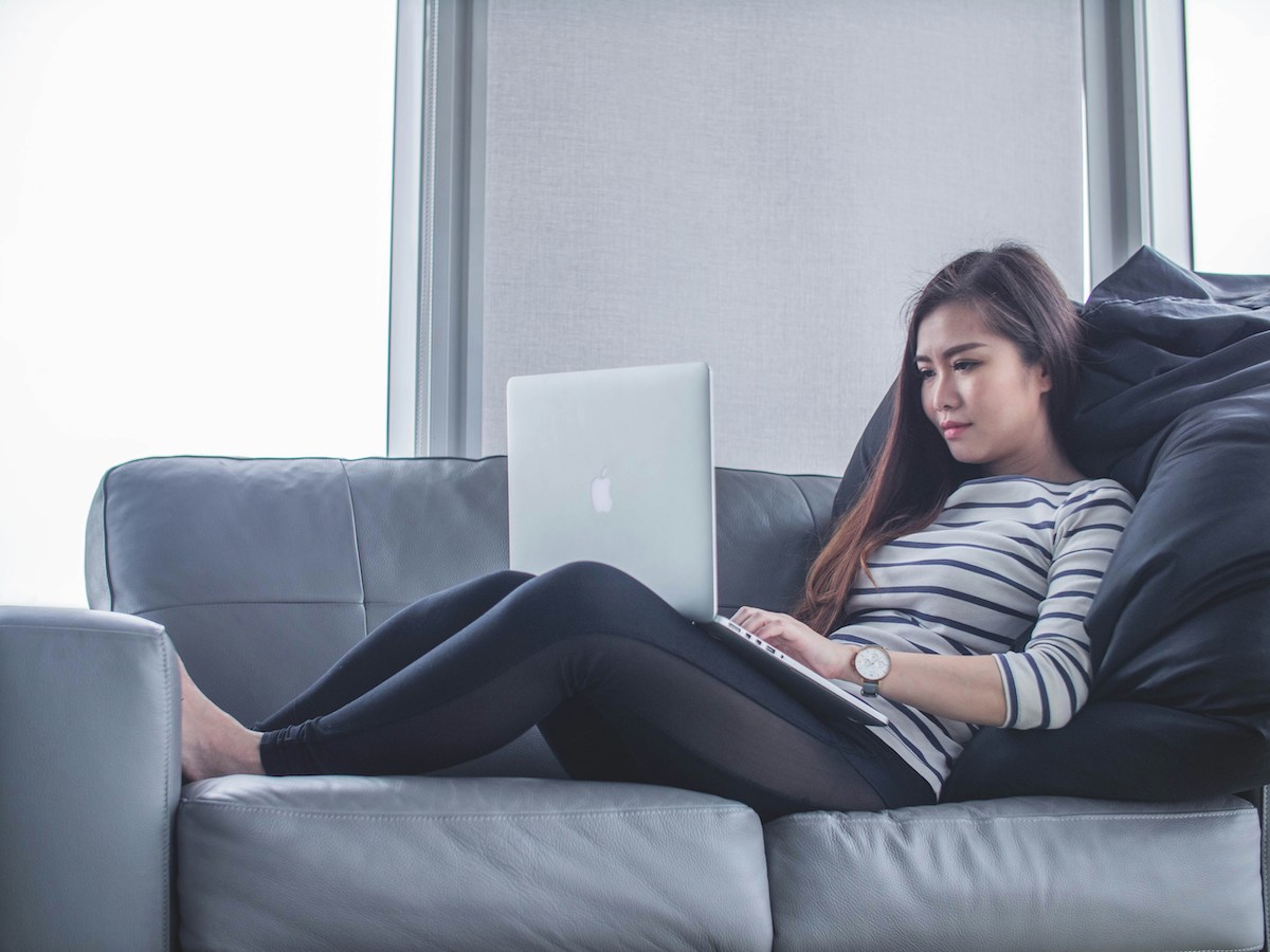 young woman with laptop on lap reading an article while laying on her couch