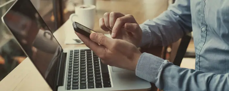 Person sitting at their laptop while scrolling on their mobile phone