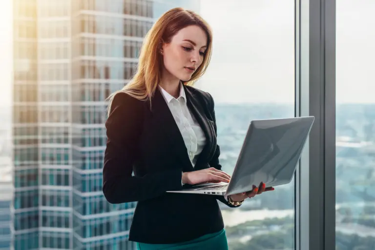 Young female lawyer working in her luxurious office holding a laptop standing against panoramic window with a view on business district