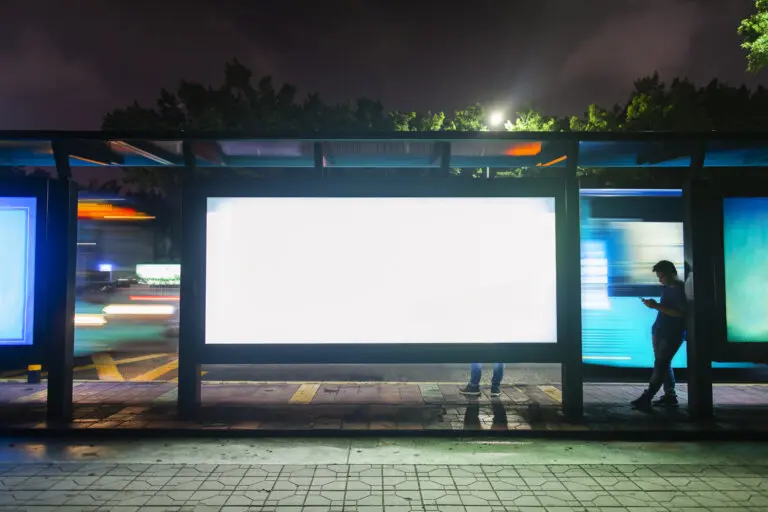 Man waiting at bus stop looks at his phone with billboard behind him.